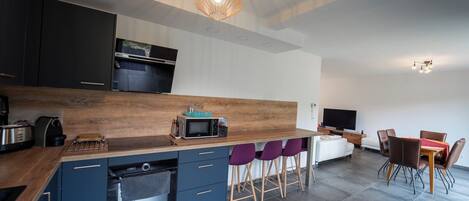 Una cocina moderna con armarios oscuros, encimera de madera y horno empotrado. Hay sillas moradas y una mesa de comedor en la zona de comedor contigua.