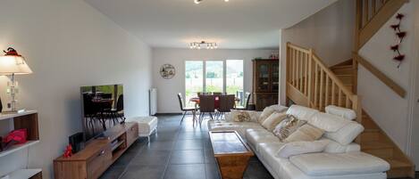 A spacious living room with a white sofa, wooden entertainment unit, and a dining area with chairs and a table. A staircase with wooden railings leads to an upper floor.
