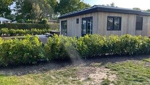 A modern, single-story building with large windows and a flat roof, surrounded by well-maintained bushes and a grassy area. - Chalet by Water in Idskenhuizen Recreation Park (Idskenhuizen)