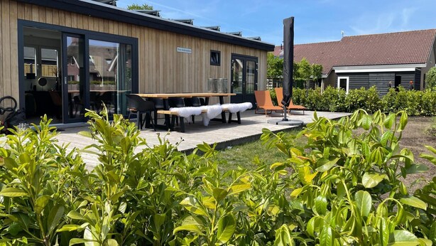 A modern building with a wooden facade, a covered outdoor seating area with tables and chairs, and a garden with green foliage. - Chalet by Water in Idskenhuizen Recreation Park (Idskenhuizen)