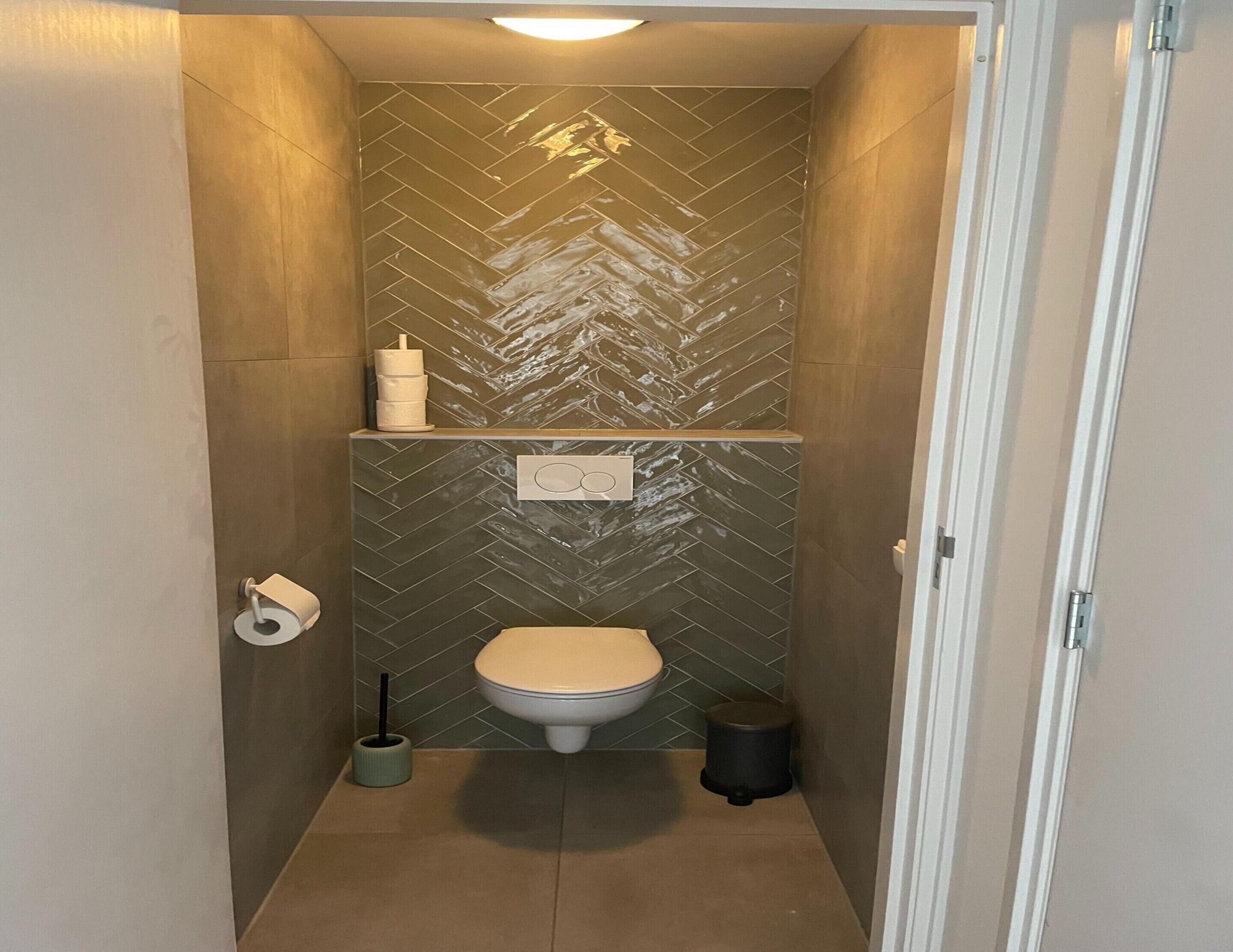 A compact bathroom with a herringbone tile backsplash, a toilet, a shelf with toiletries, and a floor-mounted bidet.