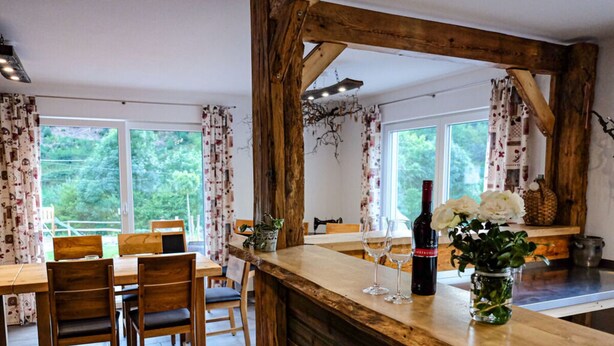 A rustic kitchen with wooden beams, a dining table with chairs, and a bar area with a bottle of wine and flowers. - Nice Apartment in Oberkirch (Oberkirch)