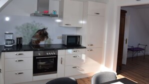 Een moderne keuken met witte kastjes, een zwarte kookplaat en een magnetron. Een kat zit op de kookplaat. Er staat een koffiezetapparaat en een klok aan de muur.