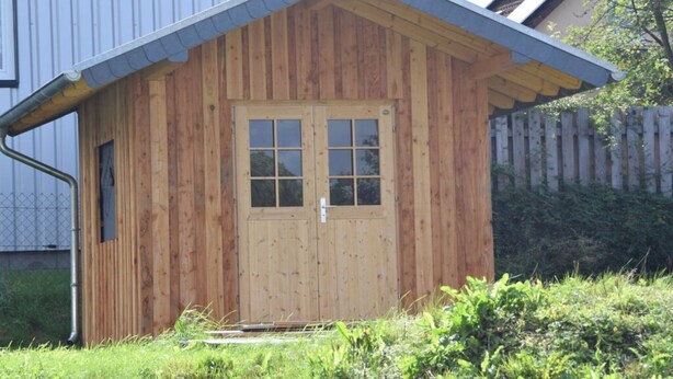 Een houten tuinhuis met een puntige dak en een dubbele deur met ruitpanelen.