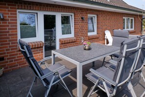 Un espace patio avec une table et des chaises, une petite plante en pot et une maison en briques dotée de fenêtres et d’une porte.