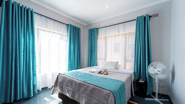 Una habitación de hotel con una cama, cortinas color turquesa, un ventilador blanco y una alfombra a rayas.