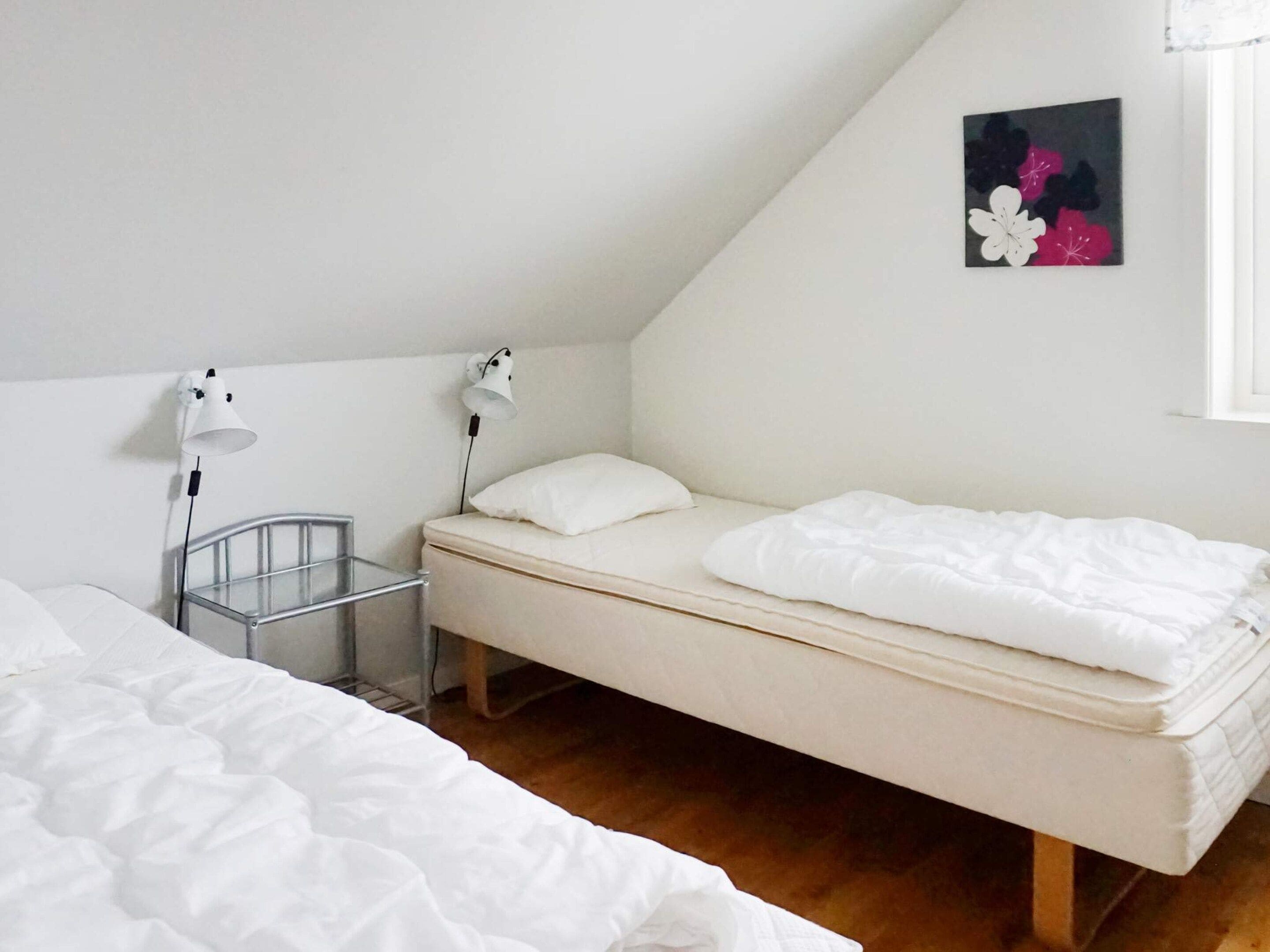 A single bed with a white mattress and pillow, a bedside table with a lamp, and a small metal rack.
