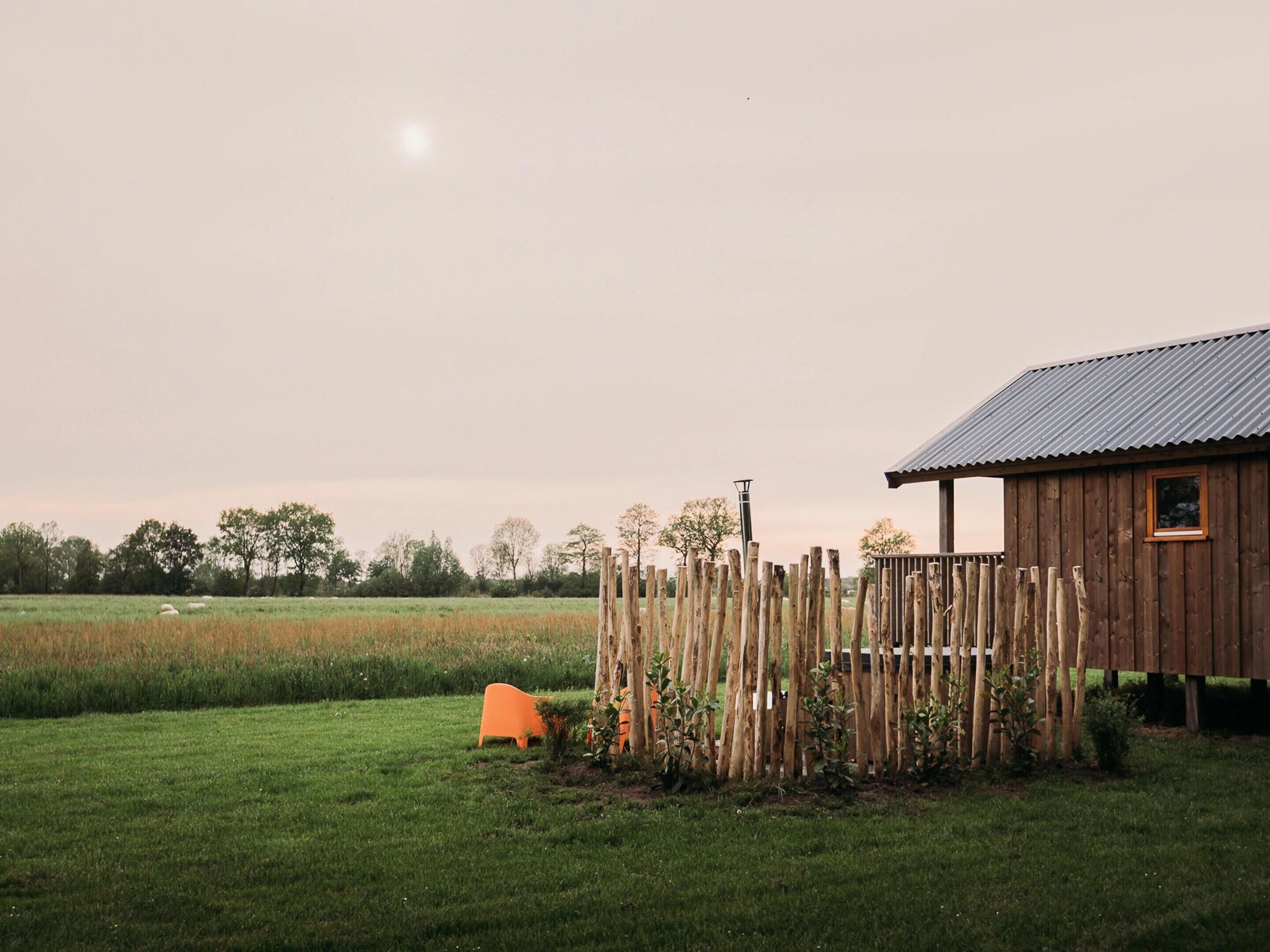 Een houten schuur met een golfplaten dak, omgeven door een grasveld en een houten hek.