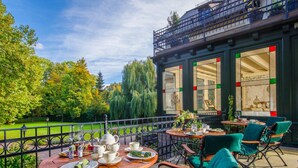 Un café en plein air avec des tables et des chaises rondes, une vue sur un jardin, et un bâtiment doté de grandes fenêtres et d’un balcon.