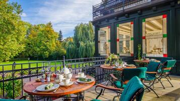 An outdoor café with round tables and chairs, a view of a garden, and a building with large windows and a balcony.