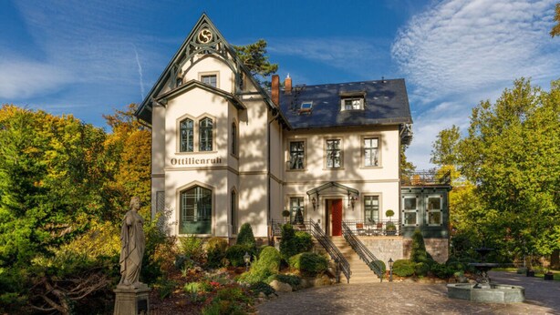 Un bâtiment imposant, doté d’une entrée remarquable, d’un escalier menant à la porte principale et d’un jardin bien entretenu, agrémenté de diverses plantes et d’une statue.
