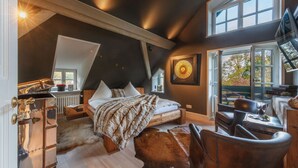 Une chambre avec un plafond en pente, un grand lit, un tapis en peau de vache, une vieille malle et une vue sur l’extérieur par une fenêtre.