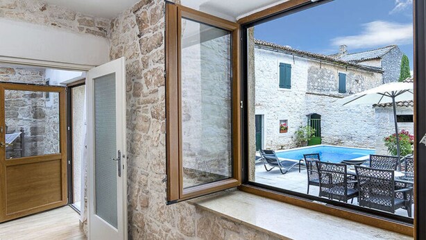 A room with a stone wall, a large window with a view of a pool and outdoor seating, and a door leading to another room. - Villa San Vincenti (Svetvincenat)