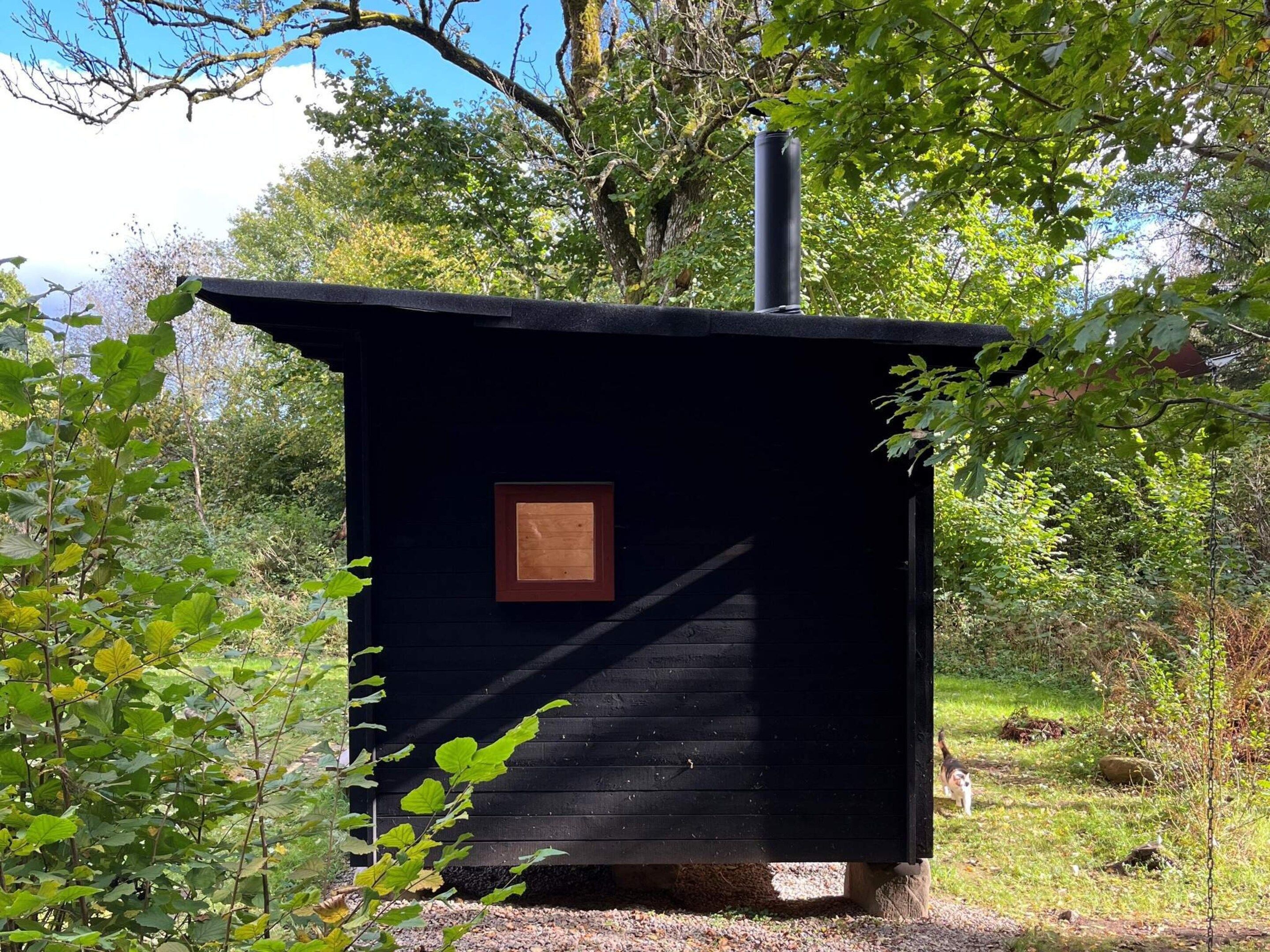 A small, dark wooden cabin with a chimney, surrounded by trees and greenery.