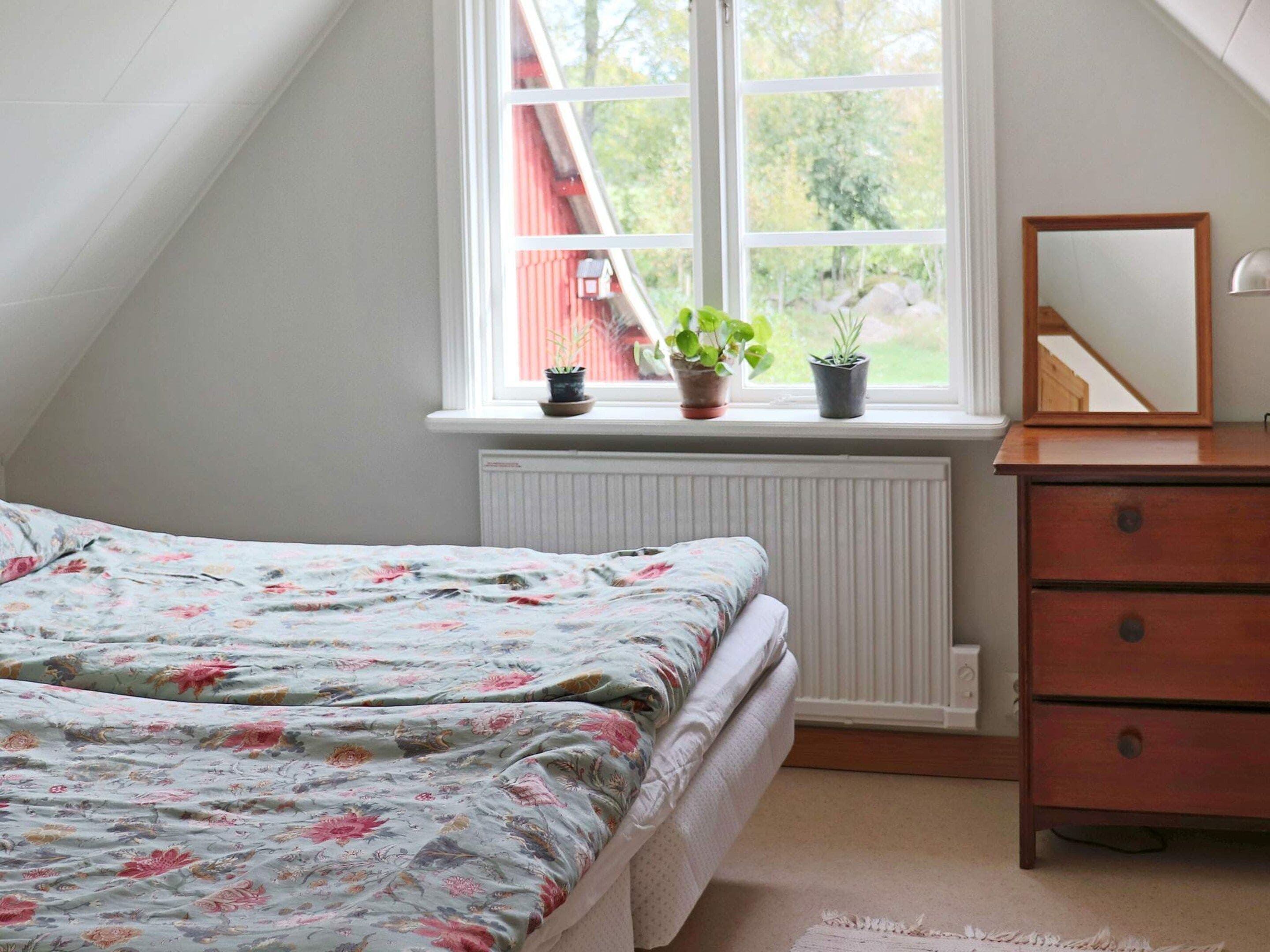 Un dormitorio con una cama, un aparador, un espejo y una ventana con plantas.