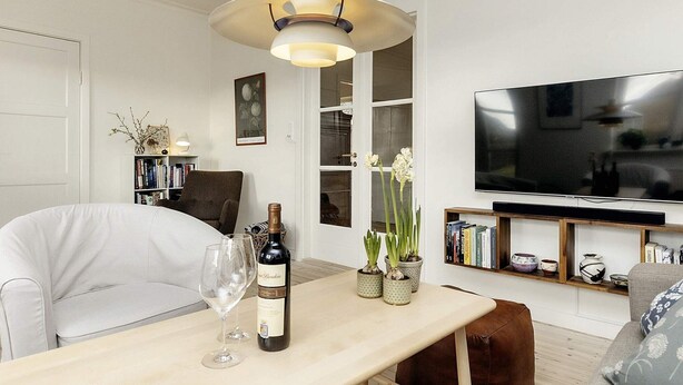 Una sala de estar moderna con una mesa de comedor de madera, un sofá blanco, un sillón marrón, un televisor y una estantería.