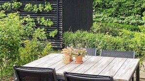 Una mesa de madera con sillas, rodeada de plantas en macetas y follaje.