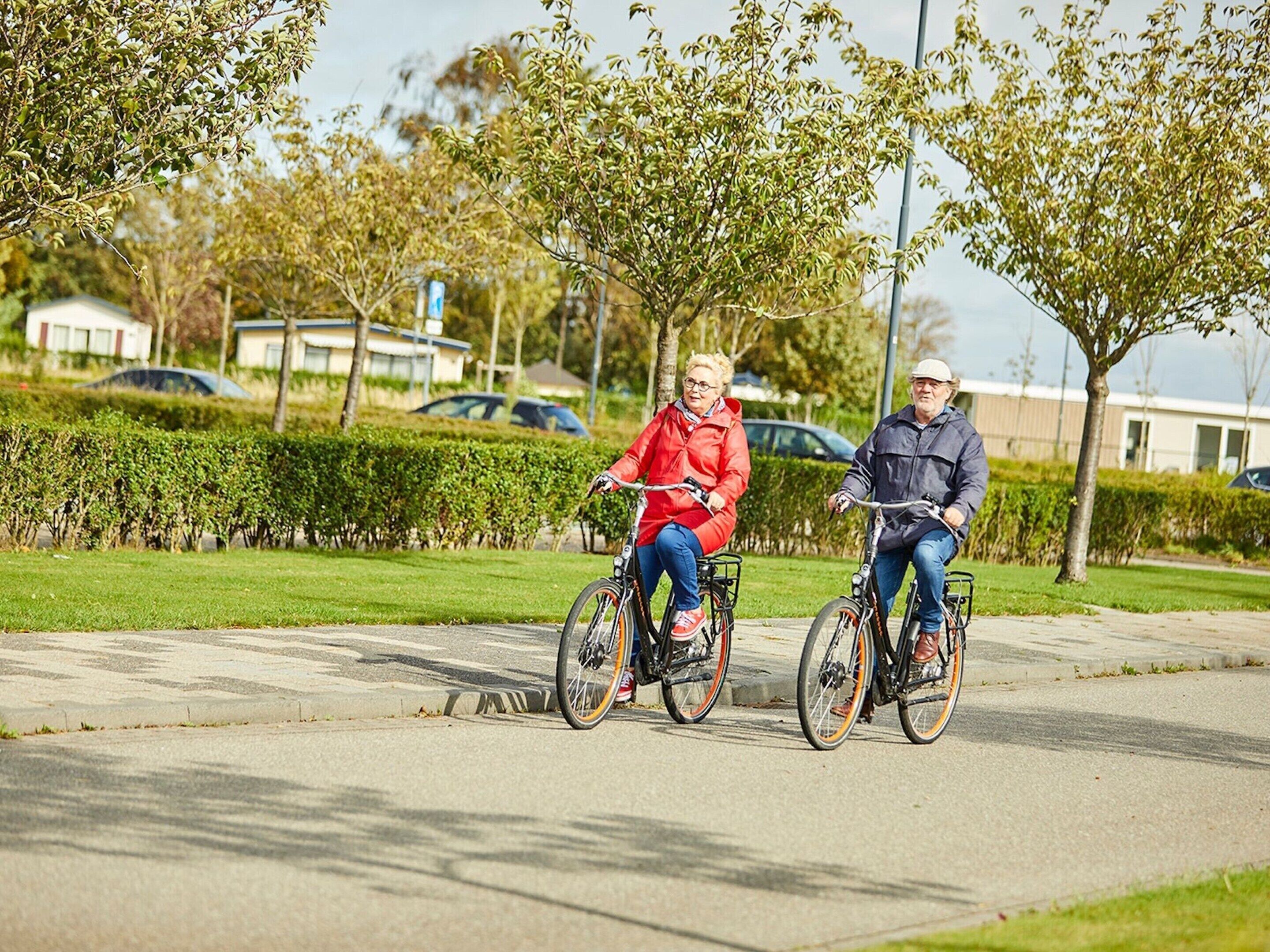 Two people riding bicycles on a paved path in a park-like setting with greenery and buildings in the background.