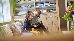 Ein gemütliches Wohnzimmer mit einer Holzhülle voller Bücher und Zeitschriften, einer bequemen Couch und einer Vase mit Blumen.