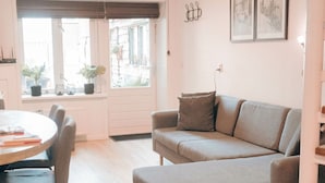Una sala acogedora con un sofá gris, una mesa redonda de madera y una ventana con plantas en macetas.