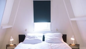 Una cama doble con ropa de cama y almohadas blancas, dos mesitas de noche con lámparas, una ventana con persiana oscura y un techo inclinado.