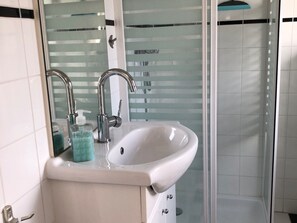 Une salle de bain avec un lavabo blanc, deux robinets, un distributeur de savon et une paroi de douche en verre.
