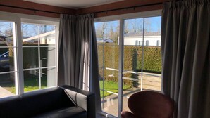 A room with a black sofa and a red chair, large windows with curtains, and a view of a garden and neighboring houses. - Tranquil Chalet Near the Wadden Sea (Tzummarum)