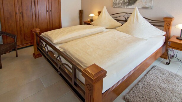 A bedroom with a wooden bed, two bedside tables with lamps, a wooden wardrobe, and a decorative wall piece. - Lovely Apartment in Hage (Berumbur)