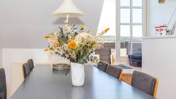 A dining area with a long table, chairs, a vase with flowers, a hanging light, and a view through a window.