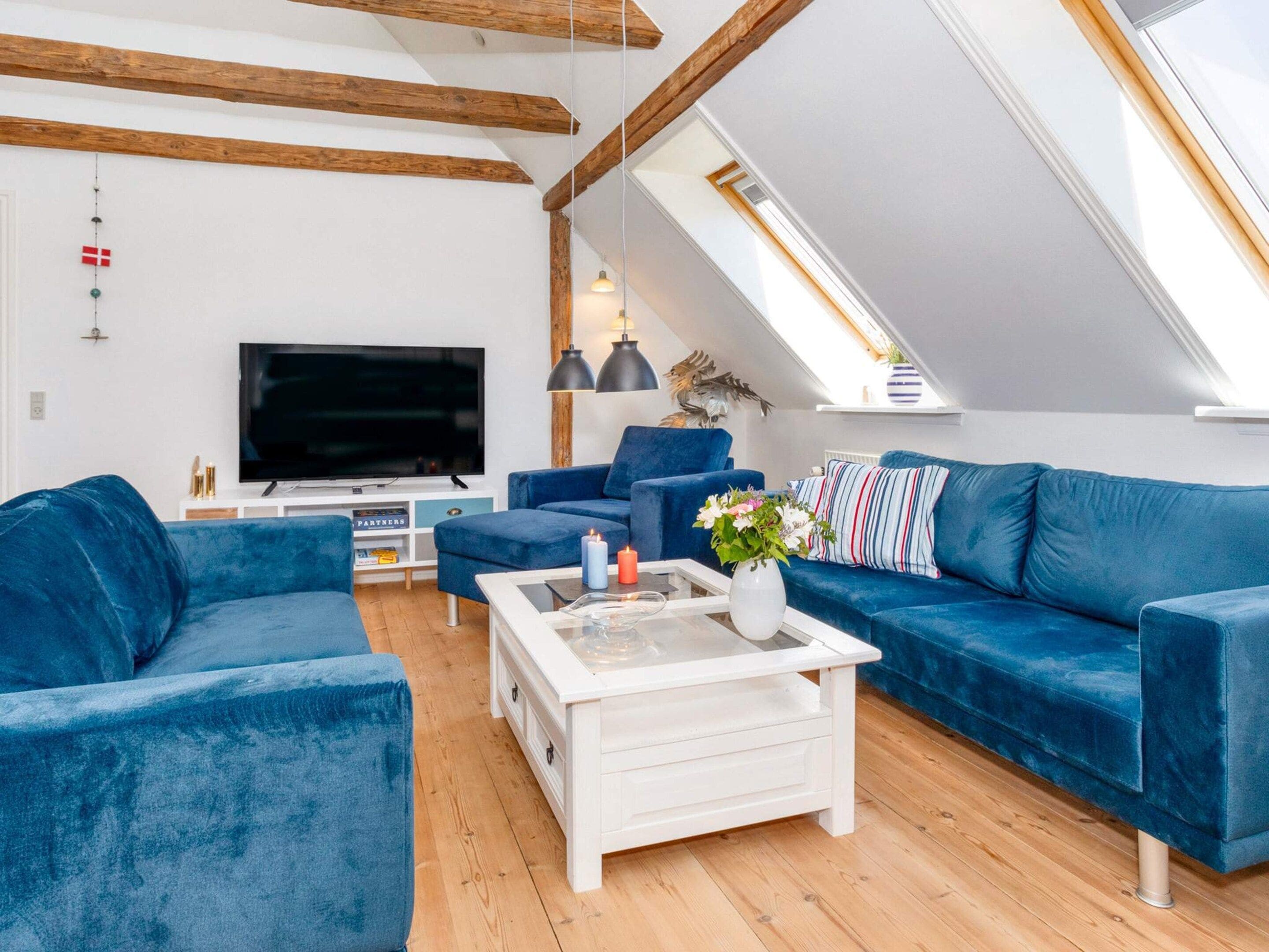 Una sala de estar moderna estilo loft con techo inclinado, vigas de madera, un televisor de pantalla plana grande, sofás azules, una mesa de centro blanca y un tragaluz.
