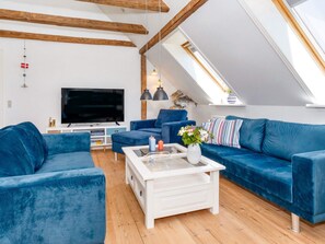 Ett modernt vardagsrum i loftstil med ett sluttande tak, träbjälkar, en stor platt-TV, blå soffor, ett vitt soffbord och ett takfönster.