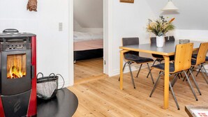 A cozy living space with a wood-burning stove, a dining table with chairs, and a bed in the background. - 4 Star Holiday Home in Thisted (Thisted)
