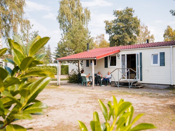 A small, single-story house with a red-tiled roof and a covered patio area. There are three people seated at a table on the patio. - Chalet in Leukermeer by the Waterside (Well)