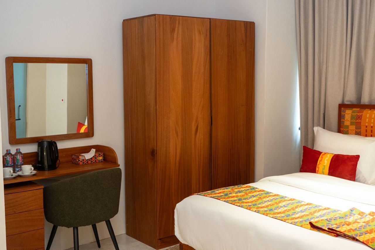 Una habitación de hotel con un armario de madera, un escritorio con silla, una cama con colcha de colores y un espejo grande en la pared.