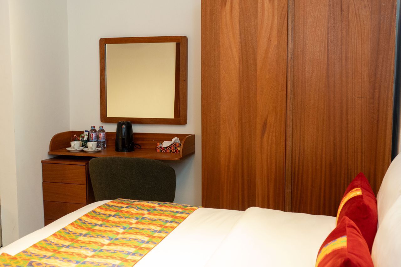 A hotel room with a bed, a wooden desk with a mirror, a chair, and a small table with a coffee maker and cups.