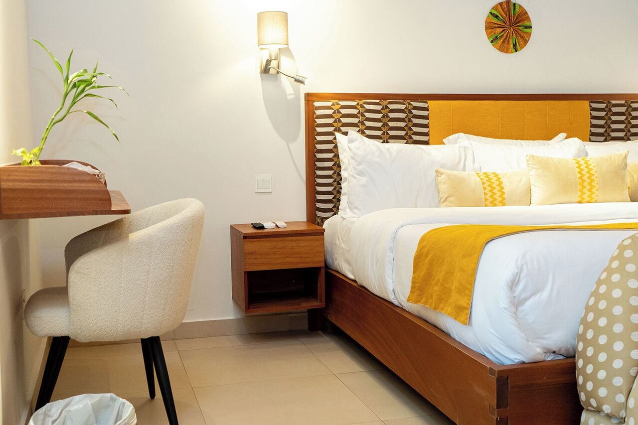 A hotel room with a wooden bed, white bedding, a nightstand, a chair, a wall-mounted lamp, and a wall art piece.