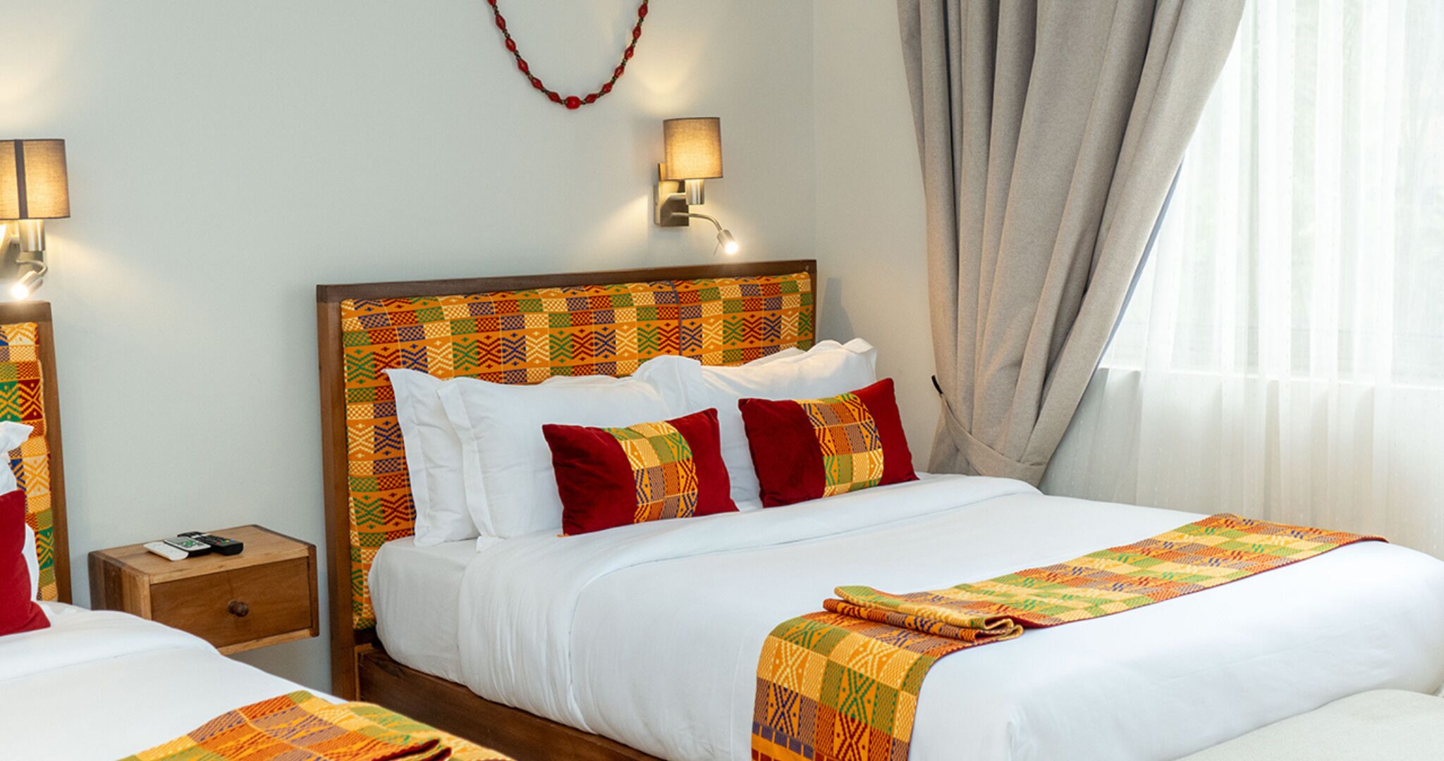 A hotel room with two beds, a wooden headboard, white bedding, red and orange patterned pillows, a nightstand with a lamp, and a window with curtains.