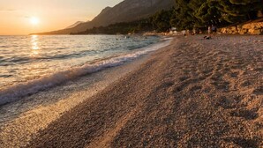Una playa de guijarros con un atardecer sobre el mar, montañas al fondo y árboles a lo largo de la orilla.