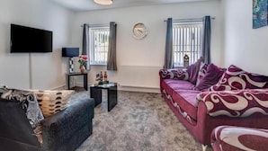 A living room with a sofa, a television, a clock, and a window with curtains. - Bay Retreat Apartment in Skinningrove (Saltburn-by-the-Sea)