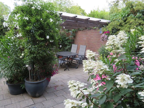 A patio area with a table and chairs, surrounded by potted plants and flowering shrubs.