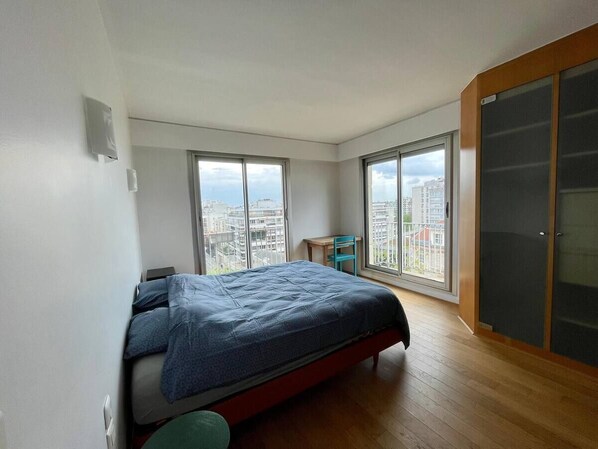 A bedroom with a bed, a desk, and large windows with a city view.