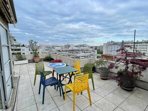 A rooftop terrace with a round table and colorful chairs, potted plants, and a view of the cityscape.