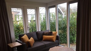 A modern living room with a dark sofa, two mustard-colored cushions, a wooden coffee table, and a small side table. Large windows offer a view of greenery outside. - Chalet With Dishwasher in a Park (Lathum)