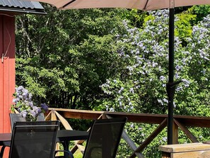 An outdoor seating area with a wooden deck, a red building, and a view of trees and purple flowers. - 4 Star Holiday Home in Annerstad (Annerstad)