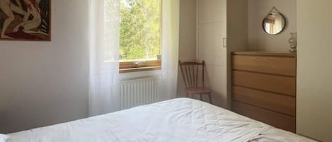 Et soveværelse med en seng, en trækommode, en stol, et vindue og et billede på væggen.