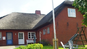 A red brick house with a thatched roof, a white door, and a small garden with green plants and two plastic chairs. - Holiday Apartment Lugworm (Ockholm)