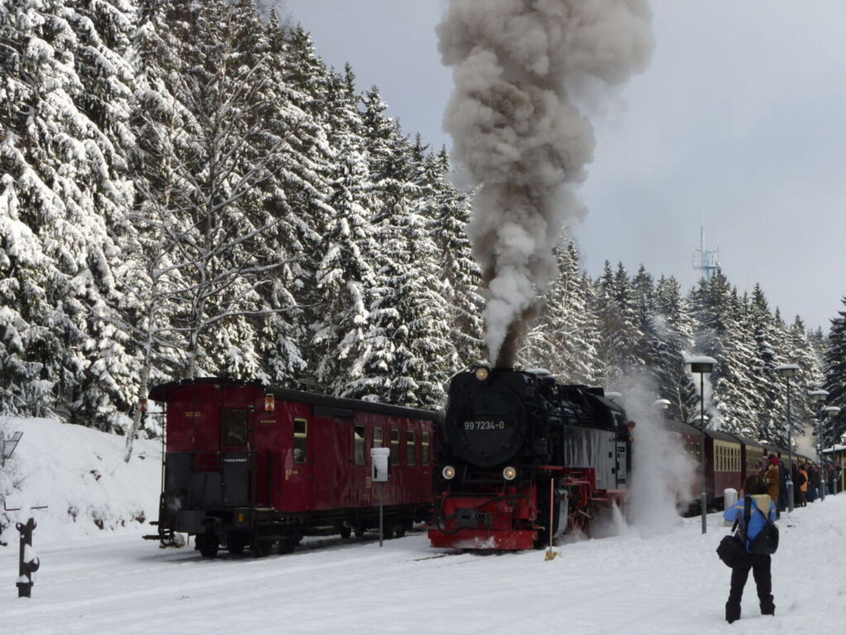 A steam locomotive with the number 99 7234-0 pulling a red passenger car through a snowy landscape with dense coniferous trees.