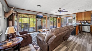 A spacious living area with a large leather sofa, a coffee table, and a fireplace. There is a kitchen area with wooden cabinets and a dishwasher. The room has large windows and a ceiling fan. - Lakefront Mtn Retreat w Private Boat Dock & Launch (Sevierville)
