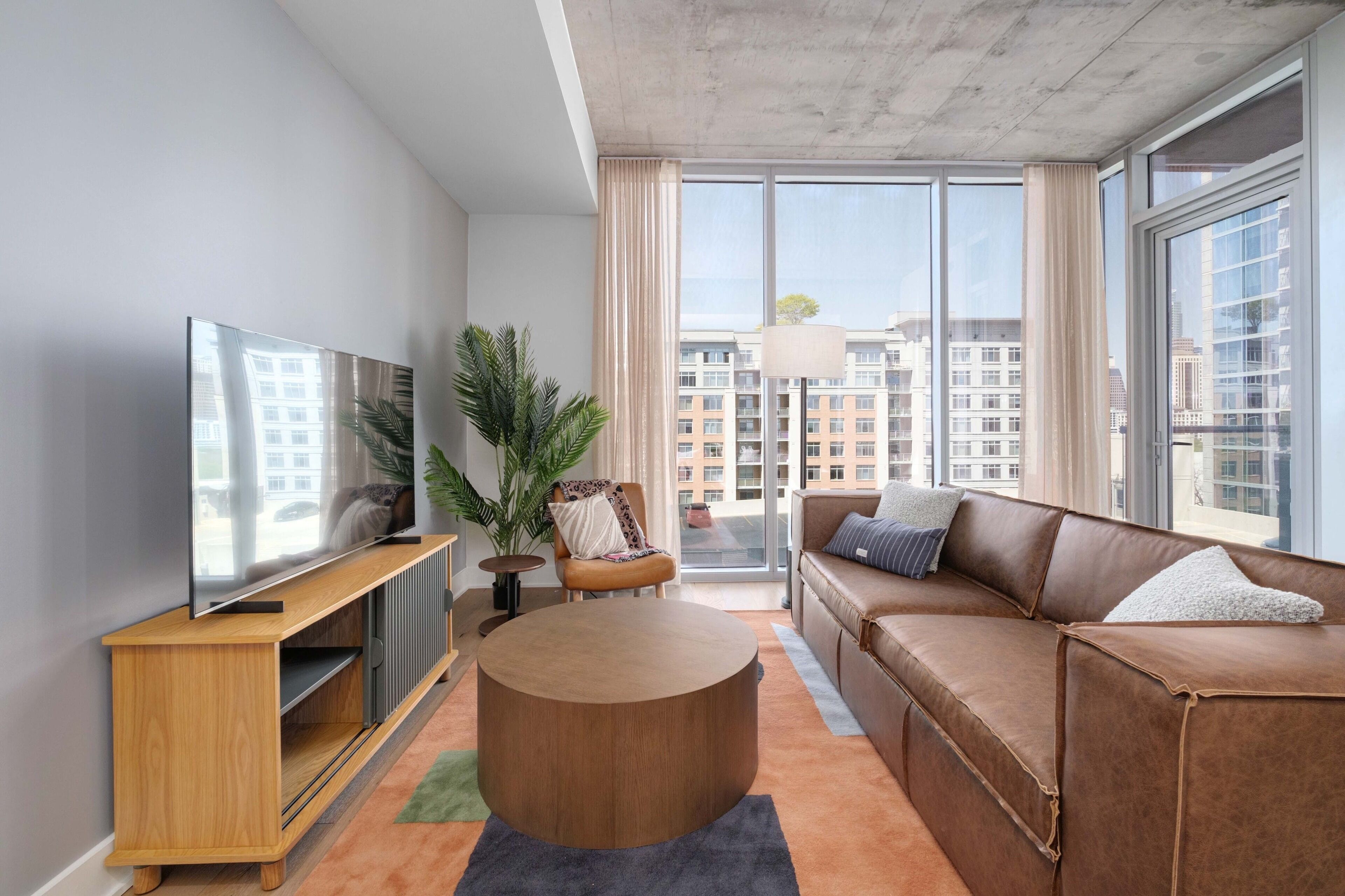 Ein modernes Wohnzimmer mit einem großen Flachbildfernseher, einem runden Couchtisch, einem braunen Ledersofa und einem großen Fenster mit Blick auf die Stadt.