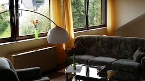 A living room with a sofa, a coffee table, and a lamp. There are windows with curtains and a view of greenery outside.